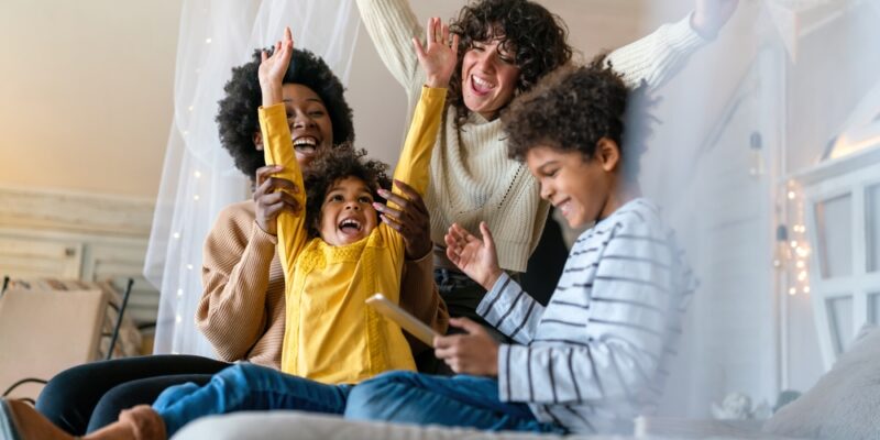 A lesbian couple excitedly plays with their children on the bed